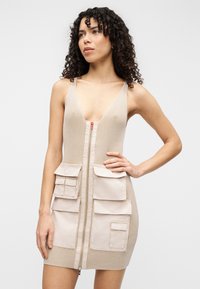 Μπεζ πλεκτό φόρεμα με βαθύ V-λαιμόκοψη, εμπρός κλείσιμο με φερμουάρ και τέσσερις τσέπες μπροστά. Το υλικό παρουσιάζει ριγωτή υφή.