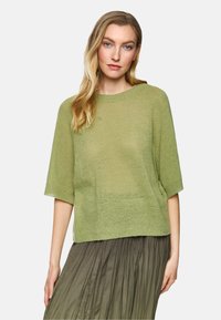 Green knit sweater with short sleeves, relaxed fit, and a ribbed crew neckline. Worn over a pleated, long olive skirt.