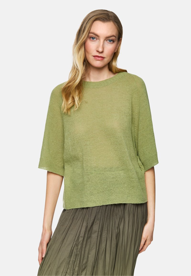 Green knit sweater with short sleeves, relaxed fit, and a ribbed crew neckline. Worn over a pleated, long olive skirt.