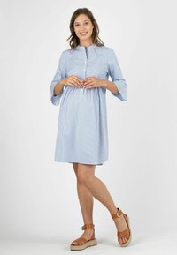 Hellblaues Hemdkleid mit vertikalen weißen Streifen, Dreiviertelärmel, geraffter Taillenbereich und schwingendem Rock. Getragen mit tanfarbigen Riemchensandalen.