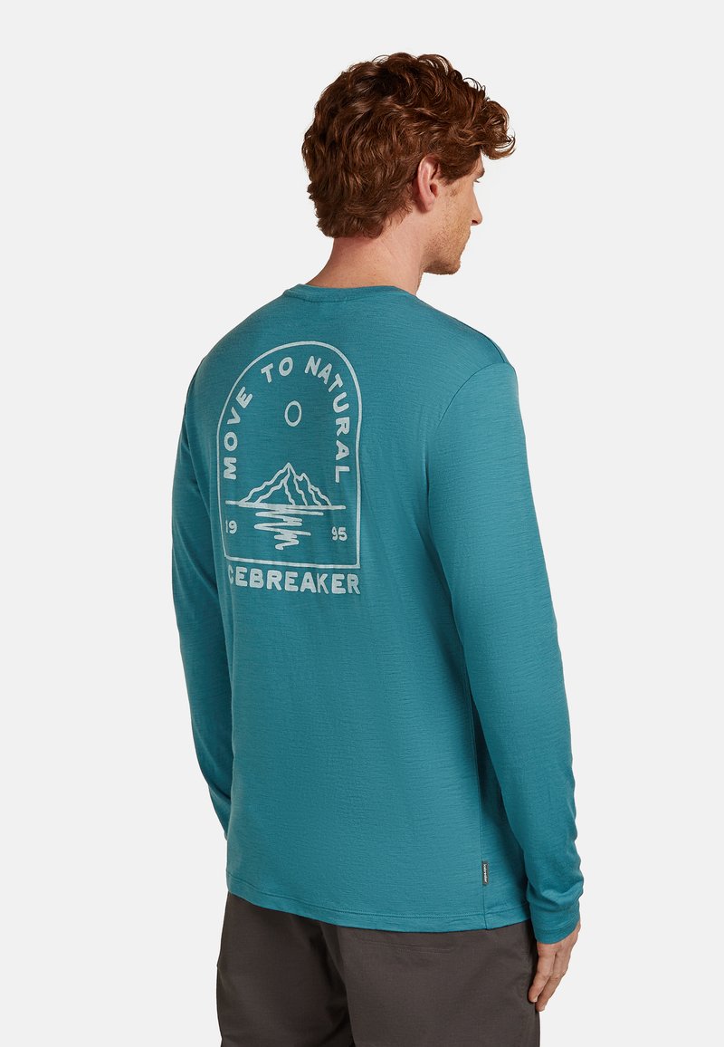 Mann mit lockigem roten Haar, der ein türkisfarbenes Langarmshirt trägt, auf dem ein Bergmotiv und der Text "Move to Natural Icebreaker" auf der Rückseite zu sehen sind.