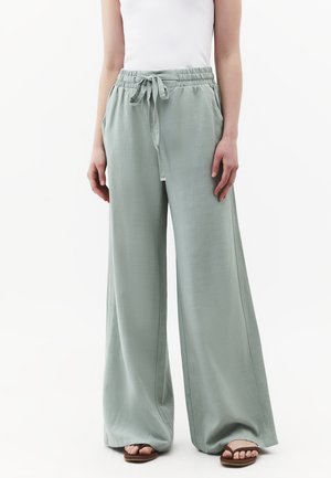 Vrouw draagt lichtgroene wijde broek met trekkoord in de taille, wit mouwloos topje en bruine sandalen, staand tegen een witte achtergrond.