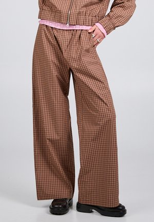 Brązowe szerokie spodnie w kratę noszone z dopasowaną marynarką i czarnymi masywnymi butami na neutralnym tle.