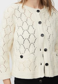 Cardigan lavorato a maglia color crema con un motivo a rombi testurizzato, bottoni neri lungo il davanti e tasche con bottone sopra pantaloni neri.