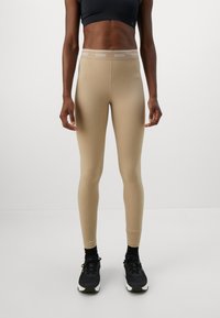 Žena u bež Guess tajicama s pojasom s logom, crnim sportskim grudnjakom, crnim čarapama i crnim Nike tenisicama, stoji protiv bijele pozadine.