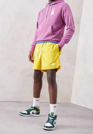Person steht und trägt einen rosa Hoodie mit Gänseblümchenmotiv, gelbe Nike-Shorts, weiße Nike-Socken und grün-weiße Nike-Sneaker.