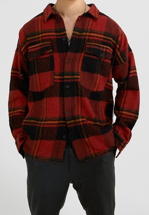 Košile s červeným a černým kostkovaným vzorem na knoflíky a s kapsami na prsou, nošená přes černé kalhoty, ruce částečně v kapsách, viditelný trup.