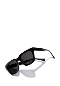 Mustad ristkülikukujulised prillid paksude raamidega ja tumedate läätsedega, mis viskavad varju valgele pinnale.