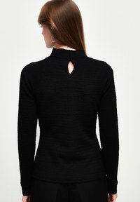 Schwarzes Langarmoberteil aus strukturiertem Stoff, mit hohem Kragen und einem Schlüsselloch-Ausschnitt am Rücken. Figurschmeichelnde Silhouette.