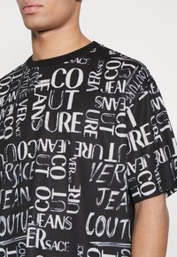 Černé tričko s krátkým rukávem s výrazným bílým textovým vzorem v různých stylech, na kterém je napsáno "VERSACE," "COUTURE" a "JEANS." Textury vypadají hladce.