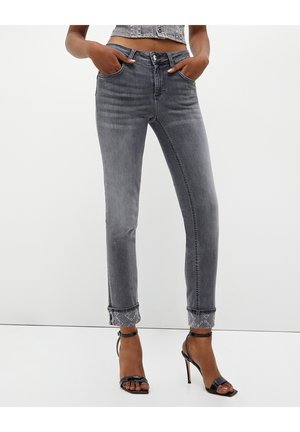 Mujer con vaqueros grises de cintura alta con puños doblados y sandalias negras de tacón alto con punta abierta, con las manos en los bolsillos.