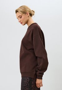 Hnedá mikina s dlhými, voľnými rukávmi a rebrovaným lemom. Tkanina sa zdá byť mäkká a štruktúrovaná. V kombinácii s vzorovanými spodnými kusmi oblečenia.