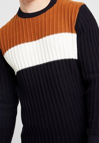 Sweter dzianinowy z poziomymi prążkami w kolorze brązowym, białym i granatowym. Model z okrągłym dekoltem, długimi rękawami i fakturowanym materiałem.