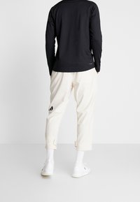 Schwarzes langärmliges Shirt mit rundem Halsausschnitt, kombiniert mit cremefarbenen, knöchellangen Hosen mit schwarzem Logo-Detail an der Seite. Weiße Sneakers.