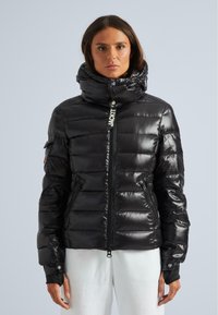 Frau, die eine schwarze glänzende Pufferjacke mit hohem Kragen und Reißverschluss-Taschen trägt, kombiniert mit weißen Hosen, steht vor einem schlichten Hintergrund.