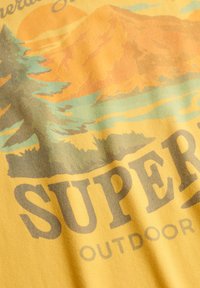 Graphique de montagne et de pin avec le texte "Superdry Outdoor" sur un tissu jaune, suggérant un thème extérieur ou nature.