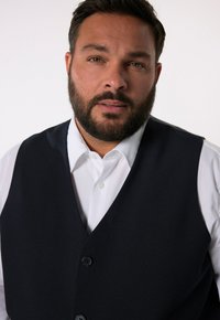 Granatowa kamizelka z teksturowanej tkaniny noszona na białej koszuli. Posiada dekolt w serek i trzy czarne guziki z przodu.