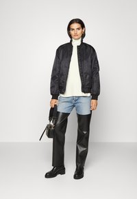 Schwarze Bomberjacke, leichter Pullover, mittelblaue Jeans, hochgeschnittene schwarze Kunstlederhosen, schwarze Plateastiefel, kleine Handtasche.