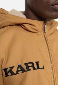 Tan hooded jacket with a zip front, featuring black embroidered "KARL" logo. Lined with soft, cream-colored sherpa texture.