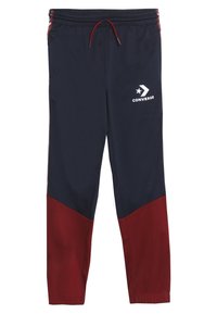 Marineblauwe en kastanjebruine sweatpants van een gladde stof. Heeft een elastische tailleband met een trekkoord en een wit Converse-logo op de dij.