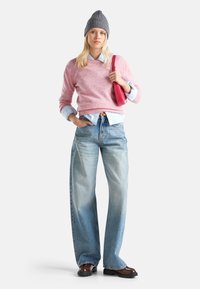 Różowy sweter dziany, jasnoniebieska koszula z guzikami, szerokie, wyblakłe niebieskie dżinsy, szary beanie, trzymająca małą różową torebkę, w brązowych butach.