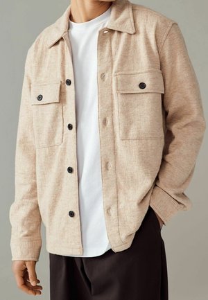 Beige wollene Hemdjacke mit Knöpfen und zwei aufgesetzten Klappentaschen vorne, getragen über einem weißen T-Shirt und kombiniert mit einer dunklen Hose.