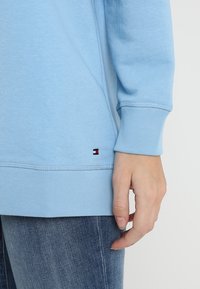 Polsini e orlo di una felpa azzurra chiara con un piccolo logo rosso, bianco e nero sopra dei jeans di denim blu e una mano rilassata.