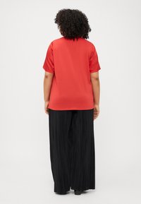 A person with curly hair wears a red t-shirt and black pleated trousers, viewed from the back against a plain white background.