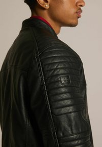 Czarna skórzana kurtka z pikowanymi detalami na ramionach, z zapięciem na zamek błyskawiczny oraz małym wytłoczonym emblemą na górnej części ramienia. Gładka tekstura.