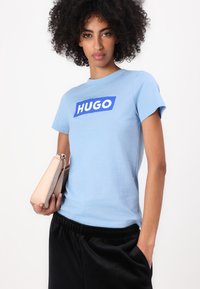 Šviesiai mėlynas medvilninis marškinėliai trumpomis rankovėmis, su ryškiai mėlynu stačiakampiu "HUGO" logotipu krūtinėje. Matosi juodos elastingos juostos kelnės.