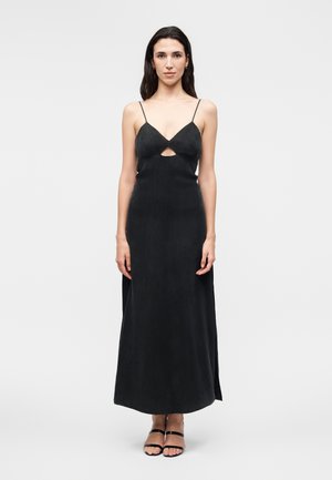 Femme debout portant une robe noire sans manches longue jusqu'à la cheville avec fines bretelles, découpe frontale, et sandales noires à lanières sur fond blanc uni.