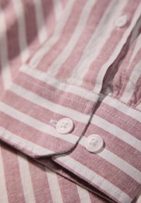 Camicia a righe in morbido tessuto rosa e bianco, con colletto button-down, due bottoni bianchi e una superficie testurizzata e tessuta.