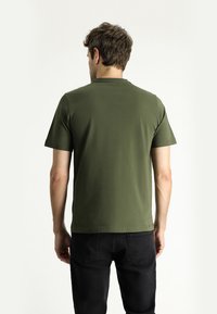 Camiseta de manga corta verde oliva en una tela suave y elástica con cuello redondo, mostrada desde atrás con jeans negros.