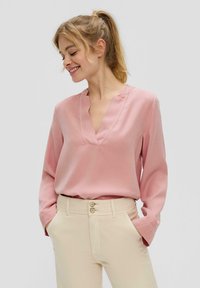 Roze blouse met lange mouwen en een V-hals, gemaakt van een gladde stof. Gecombineerd met lichtbeige broeken met een sluiting met twee knopen.