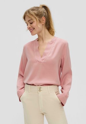 Roze blouse met lange mouwen en een V-hals, gemaakt van een gladde stof. Gecombineerd met lichtbeige broeken met een sluiting met twee knopen.