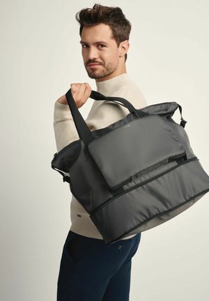 Duża czarna torba z nylonu z przednią kieszenią na suwak, podwójnymi uchwytami i lekko wyściełanym designem, noszona na jednym ramieniu przez mężczyznę.