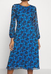 Blå midi klänning med svart blommönster, långa genomskinliga ärmar, rynkad halsringning och åtsittande midja på en stående person.