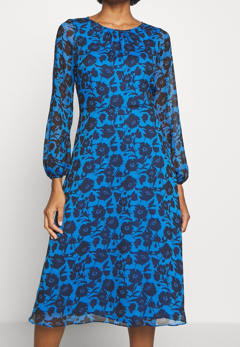 Blå midi klänning med svart blommönster, långa genomskinliga ärmar, rynkad halsringning och åtsittande midja på en stående person.