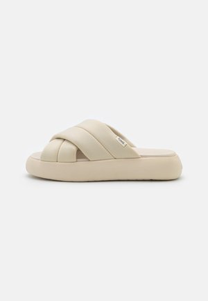 Comfort slide sandal in off-white, featuring crossed soft straps, a flat, thick sole, and a matte finish. Brand label visible on side.