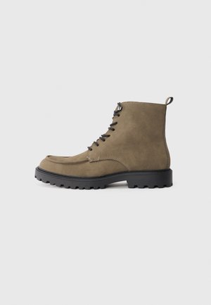 Bota de ante marrón con suela de goma negra, punta redondeada, cordones en la parte delantera y costuras en contraste. Presenta un lazo en el talón para facilitar su colocación.
