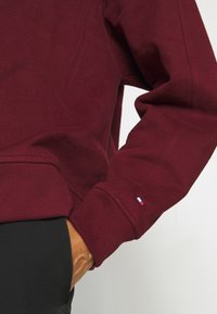 Burgundowa bluza z ribbowanymi mankietami, z małą metką z logo na rękawie. Wykonana z miękkiego materiału bawełnianego.