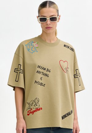 Frau trägt beiges Oversized-T-Shirt mit Phrasen und Grafiken, darunter "Dream big anything is possible," Kreuze mit der Bezeichnung "Karma Kills," und Cherubinen mit der Bezeichnung "Together."