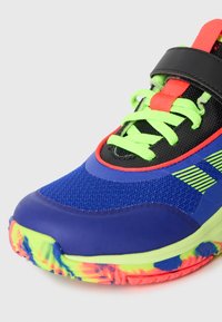 Zapatilla deportiva azul con parte superior de malla, detalles en negro, cordones verde neón y suela con patrón colorido. Cuenta con una correa de velcro para un ajuste seguro.