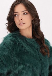 Un wrap din blană ecologică verde cu o textură moale, având un guler înalt și un design deschis în față, purtat peste un colier din lanț aurie.