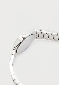 Montre en métal argenté avec un boîtier rectangulaire, des maillons de bracelet lisses et une couronne à surface texturée. Design minimaliste, finition brillante.