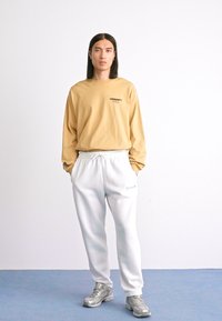 Homme avec de longs cheveux noirs portant une chemise beige à manches longues, un pantalon de survêtement blanc et des baskets argentées, se tenant avec les mains dans les poches contre un mur blanc.