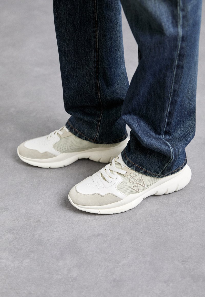 Sneakers in light gray and soft beige mesh with suede accents. Features a lace-up design and a thick white rubber sole. Worn with blue jeans.