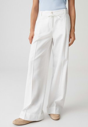 Femme portant un pantalon blanc taille haute à jambes larges avec un bouton frontal et un cordon de serrage, associé à des chaussures plates beiges.