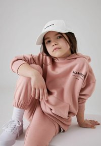 Ružičasti oversized dukserica i odgovarajuće trenerke od mekog materijala, s prednjim džepom i izvezenim logotipom. Bijela bejzbol kapa i patike.