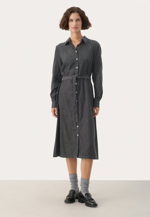 Robe boutonnée grise avec des manches longues, une taille ceinturée et une coupe droite, fabriquée dans un tissu texturé. Associée à des chaussures noires et des chaussettes grises.